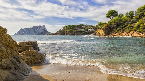 A coastal landscape with rocky shores, clear blue waters, and a distant mountain. - El Salvador Fustera (Benissa)