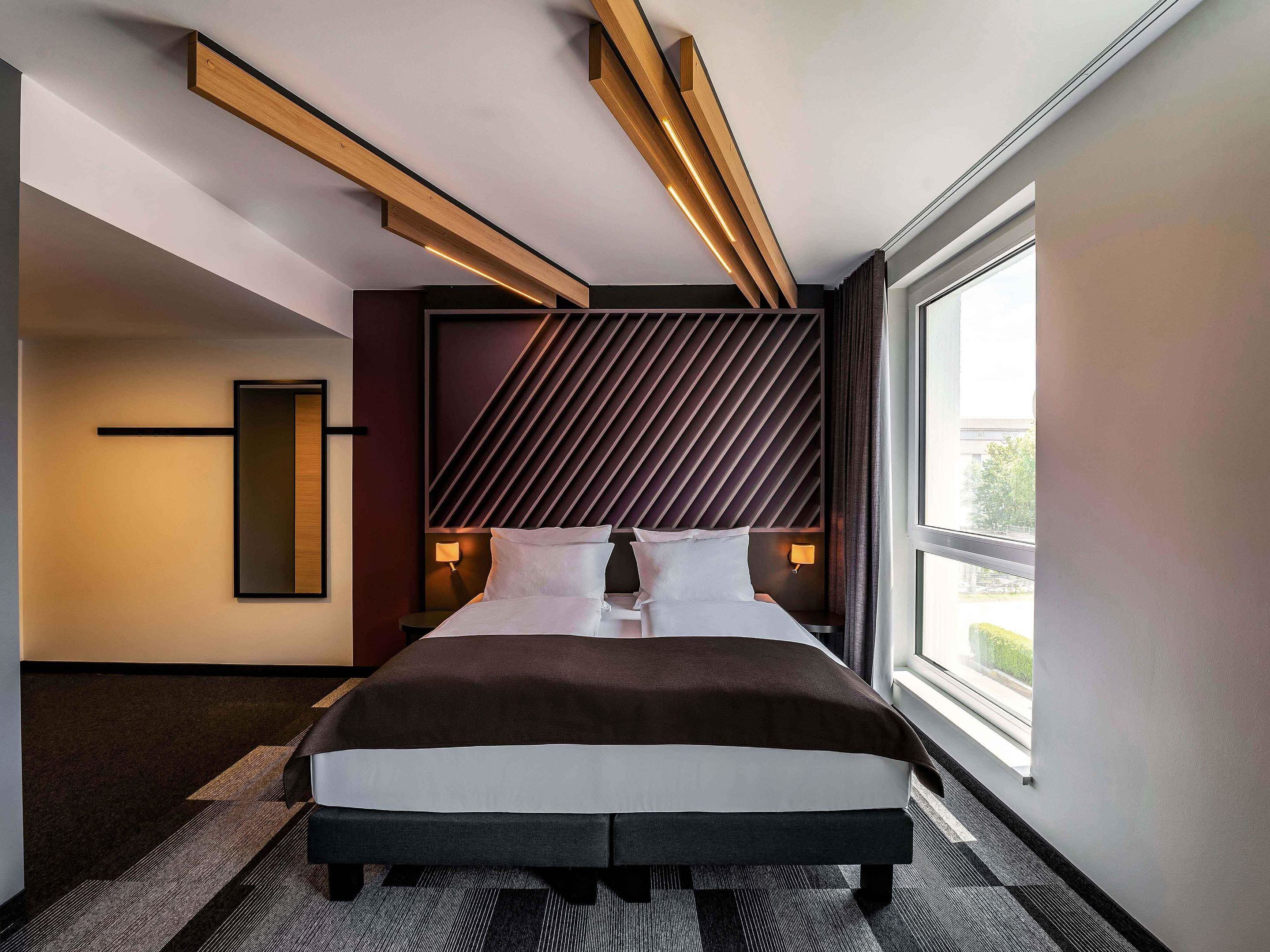 Ein modernes Hotelzimmer mit einem großen Bett, Holzbalkendecke, einer Wand mit Muster und einem großen Fenster.