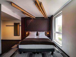 Una habitación de hotel moderna con una cama grande, vigas de techo de madera, un papel tapiz con diseño y un ventanal amplio.