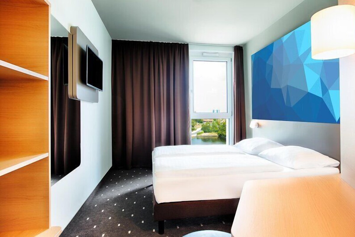 A hotel room with a bed, a TV mounted on the wall, a window with curtains, and a view of greenery outside.