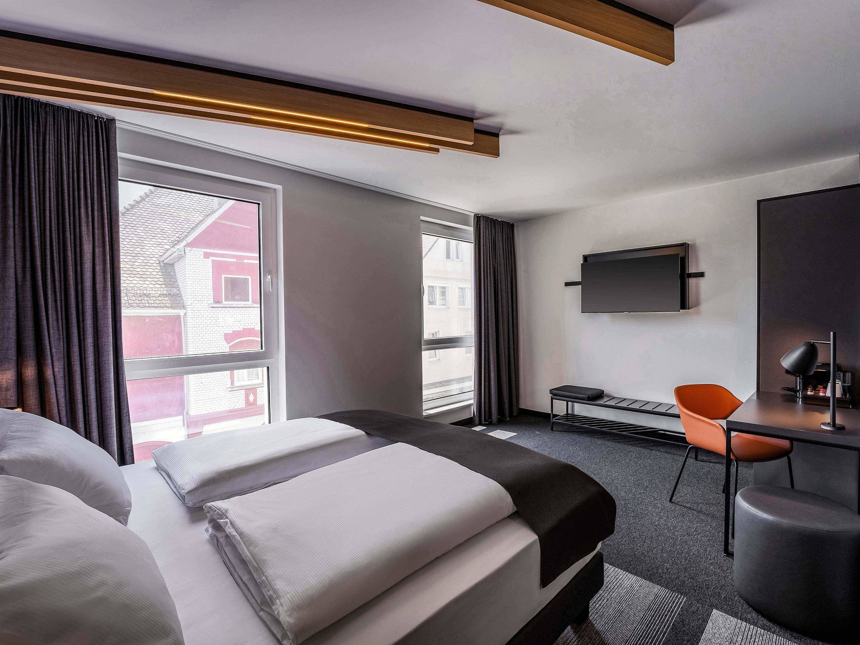 A modern hotel room with a large bed, a desk, a chair, and a TV mounted on the wall.