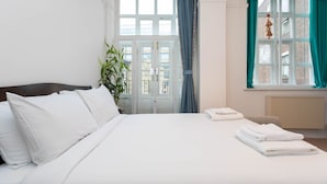 Een net opgemaakt bed met witte lakens, een potplant en een raam met blauwe gordijnen.