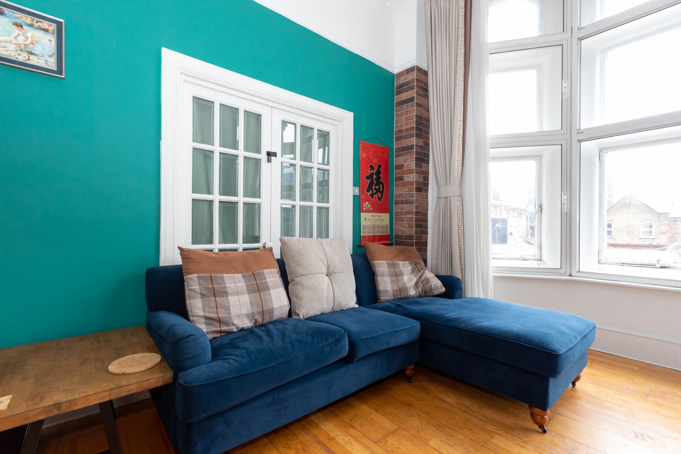 A room with teal walls, a blue sofa with cushions, a wooden table, and a window with curtains.