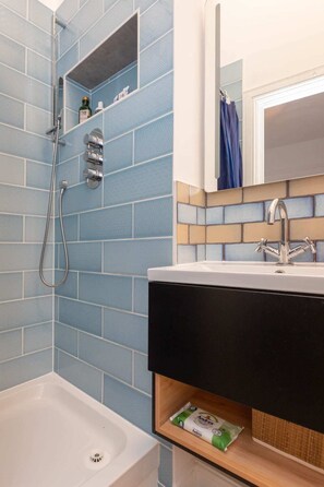 Une salle de bain équipée d’une baignoire, d’une douche avec un pommeau de douche amovible, d’un lavabo avec un miroir au-dessus et d’une étagère contenant des articles de toilette.
