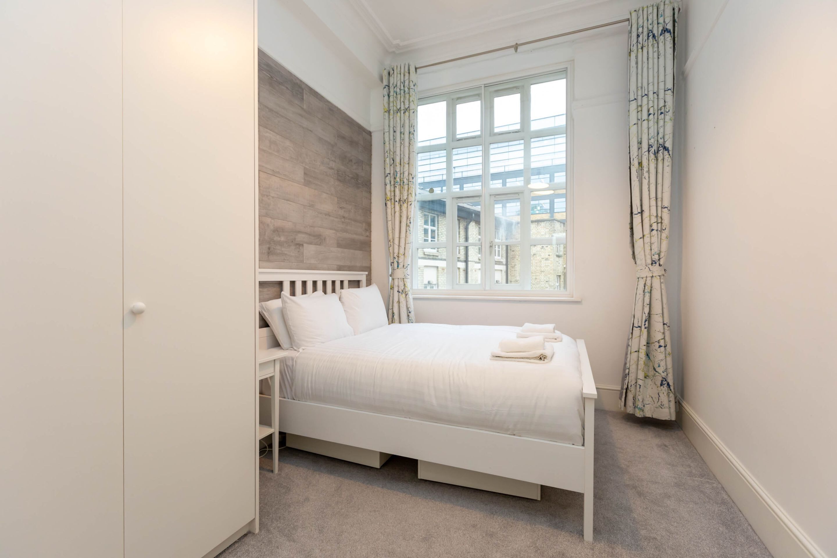 Una camera da letto con un letto bianco, una testata in legno, una finestra con tende e vista sugli edifici.