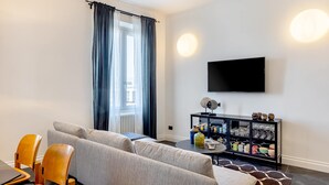 Uma sala de estar moderna com sofá, mesa de jantar com cadeiras, aparador preto com copos e uma TV de tela plana na parede.