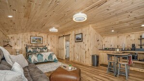 A cozy wooden cabin interior with a bed, sofa, ottoman, and a kitchen area with a bar.