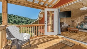 A wooden deck with a bench, a sliding glass door to a room with a sofa and a television, and a view of a forested area.