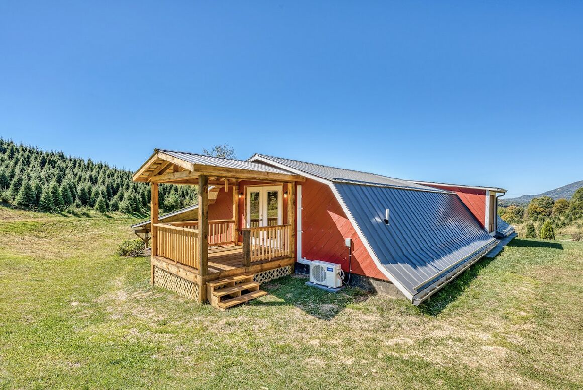 Une cabane en bois dotée d’un porche et d’un toit en pente, entourée d’un champ herbeux et d’une forêt dense.