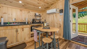 A wooden cabin interior with a kitchen area, a dining table with stools, and a sliding glass door leading to an outdoor deck.