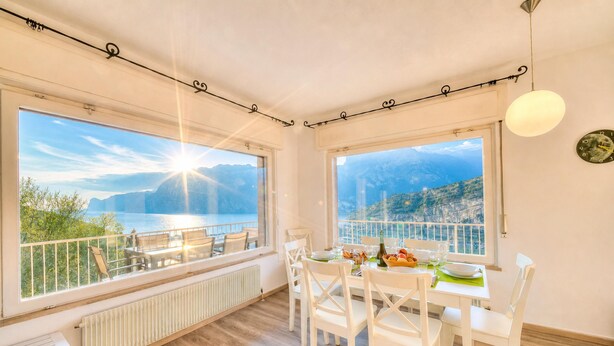 Un comedor con una mesa dispuesta para cuatro personas, ventanas amplias con vista a las montañas y al agua, y un techo blanco con una lámpara.