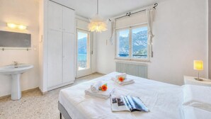 Habitación de hotel con cama, mesita de noche con lámpara, ventana con vistas a montañas, baño con lavamanos y armario.