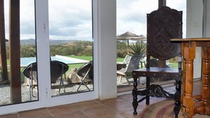 A room with a view of a green landscape through large glass doors. There is a wooden table and a chair in the room.