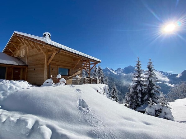 A wooden cabin with a snow-covered roof, surrounded by snow-covered trees and mountains in the background.