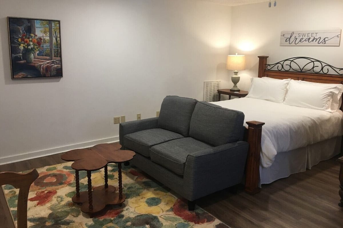 A bedroom with a bed, a gray sofa, a small table, and a chair.