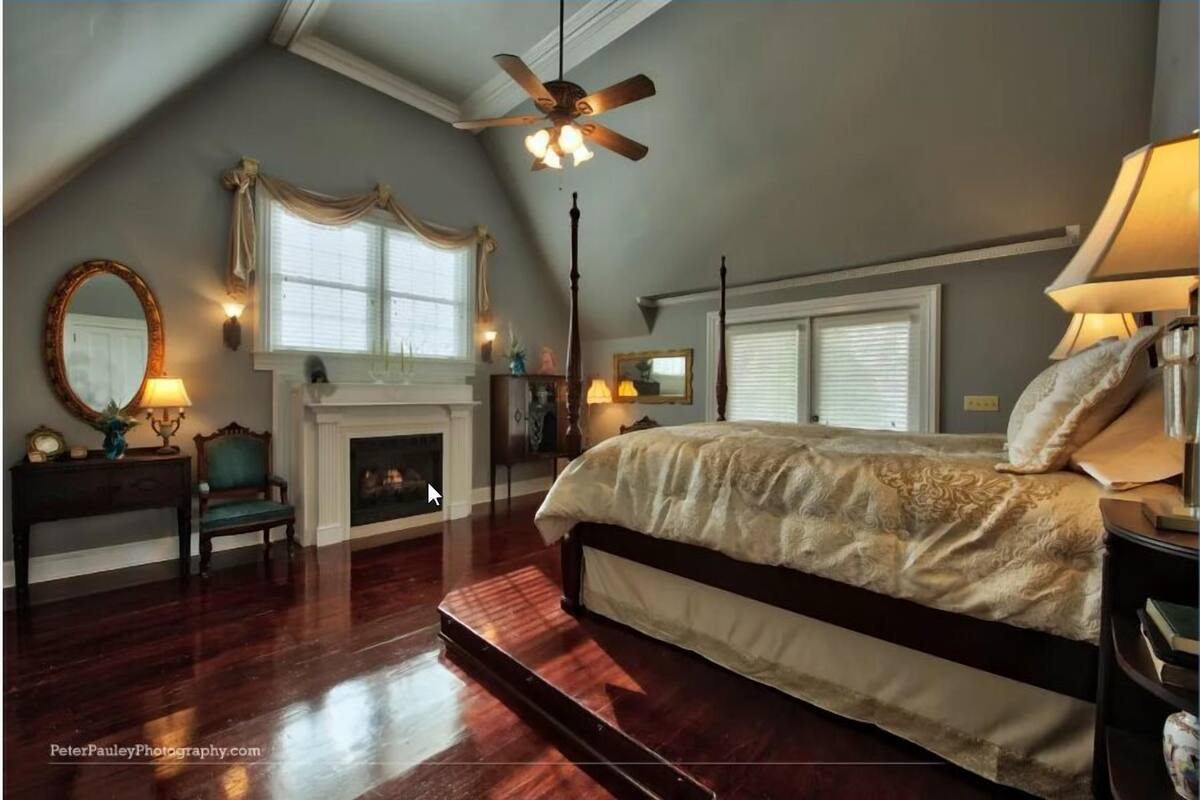 A bedroom with a large bed, a fireplace, a ceiling fan, and a view of the outdoors.