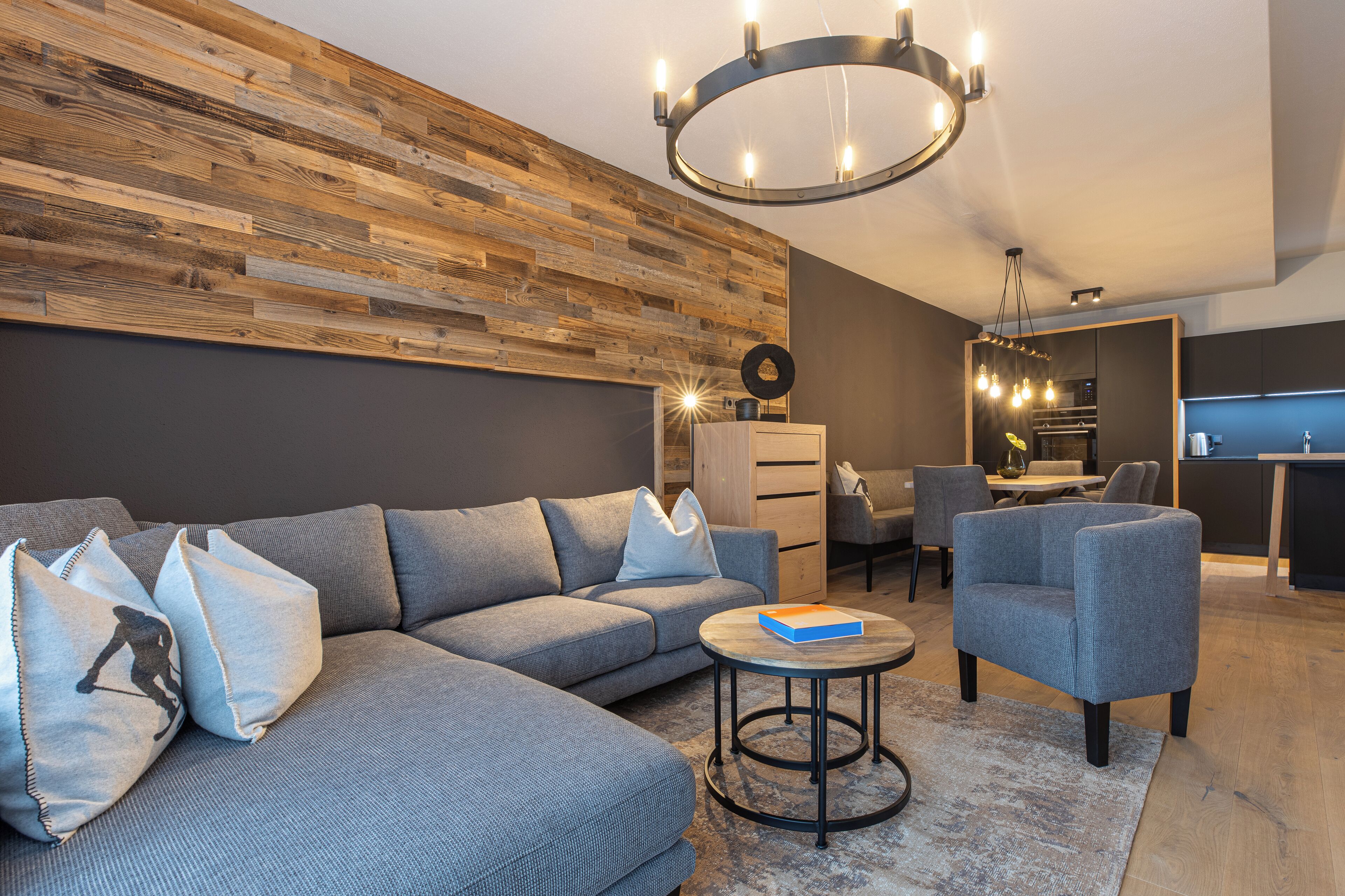 Ein modernes Wohnzimmer mit einer Eckcouch, einem runden Couchtisch und einem Essbereich, wobei im Hintergrund eine Küche zu sehen ist.