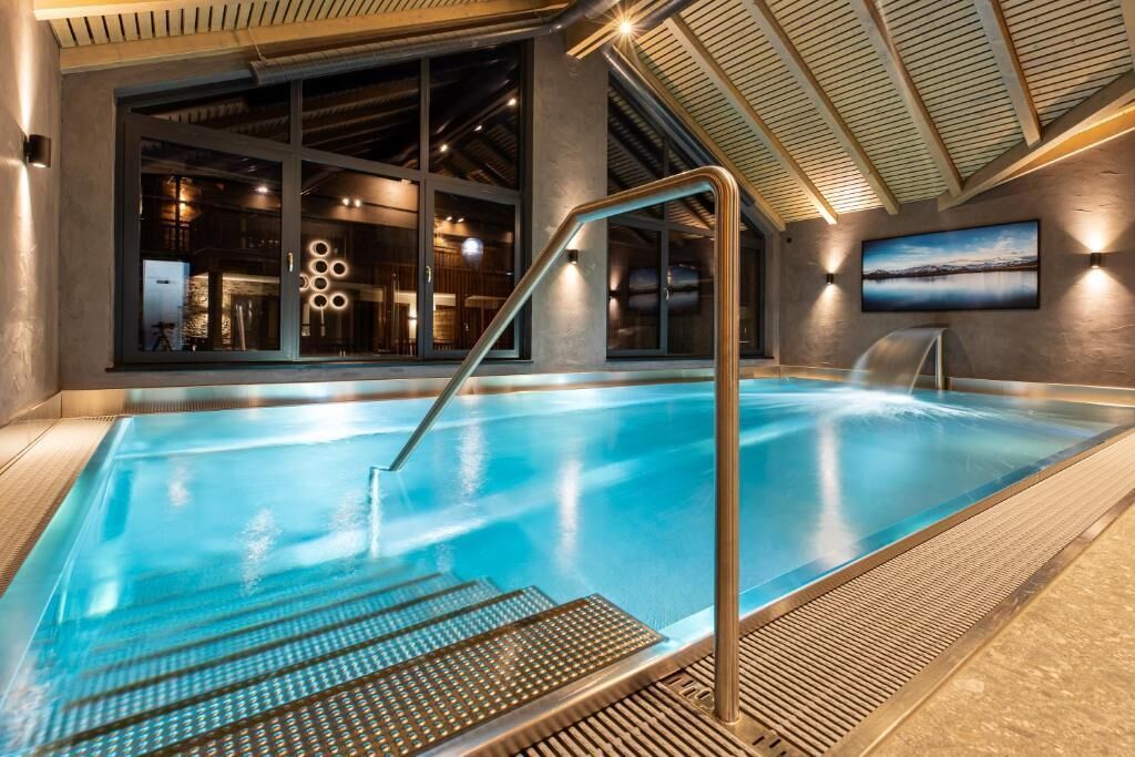 An indoor swimming pool with a clear view of the adjacent restaurant area, a metal staircase leading into the pool, and a large window providing natural light.