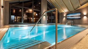 An indoor swimming pool with a clear view of the adjacent restaurant area, a metal staircase leading into the pool, and a large window providing natural light.