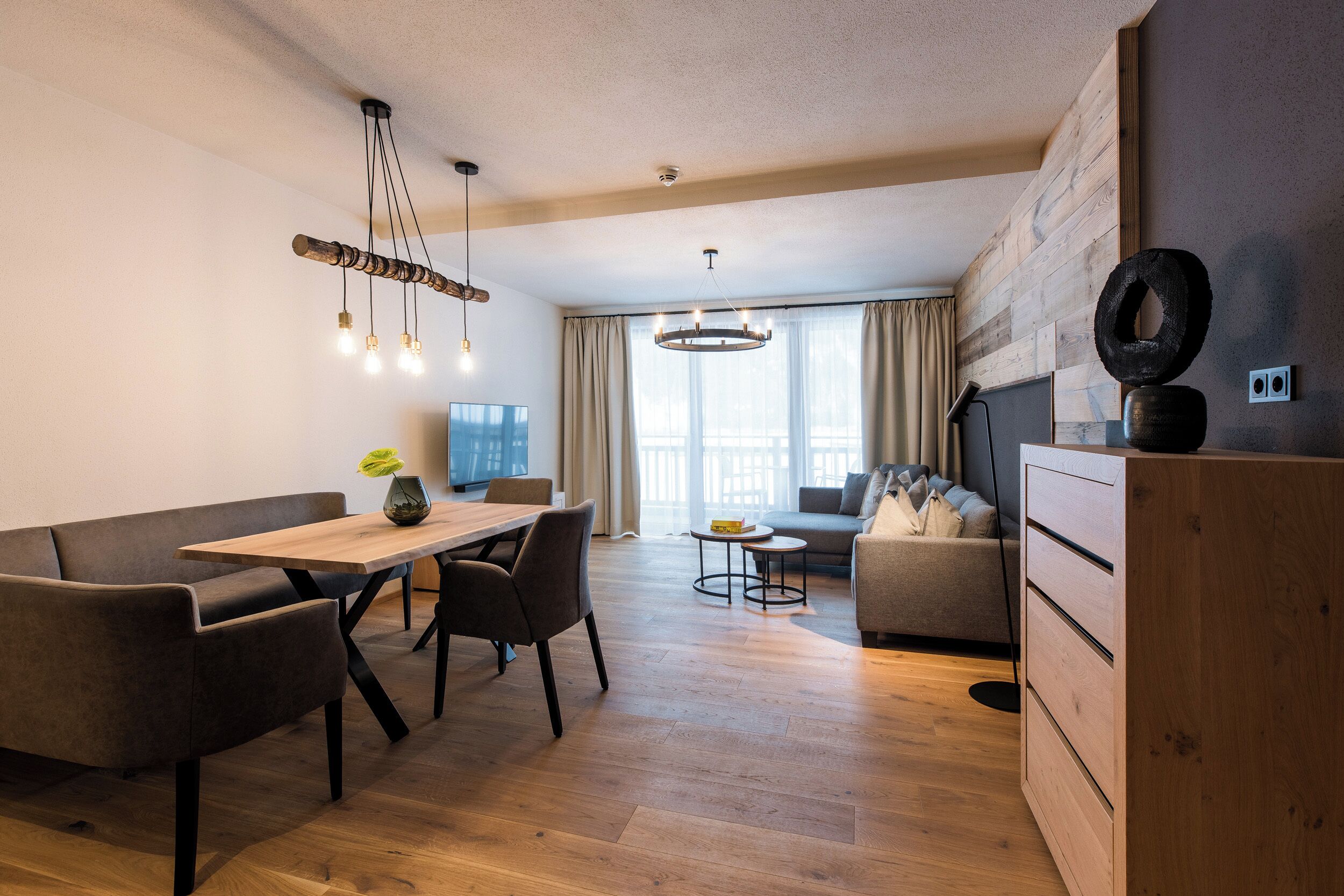 Un salon moderne avec une table à manger en bois, des canapés foncés et une grande fenêtre agrémentée de rideaux.