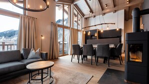 Een moderne woonkamer met een bank, een ronde tafel en een eethoek met stoelen. Er is een open haard en grote ramen met gordijnen.