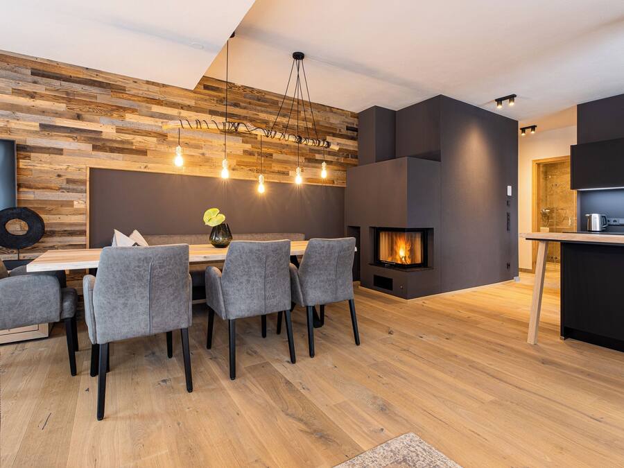 A modern dining area with a wooden wall, a fireplace, and a kitchen in the background.