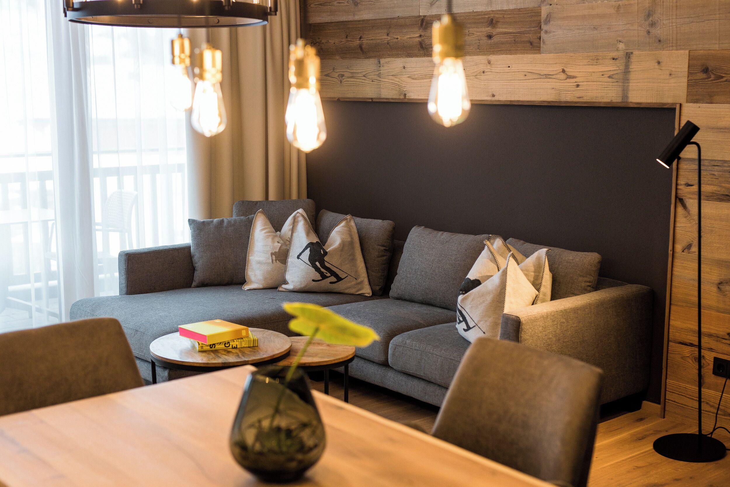 Een moderne woonkamer met een grijze bank, een ronde houten salontafel en een vaas met een plant.