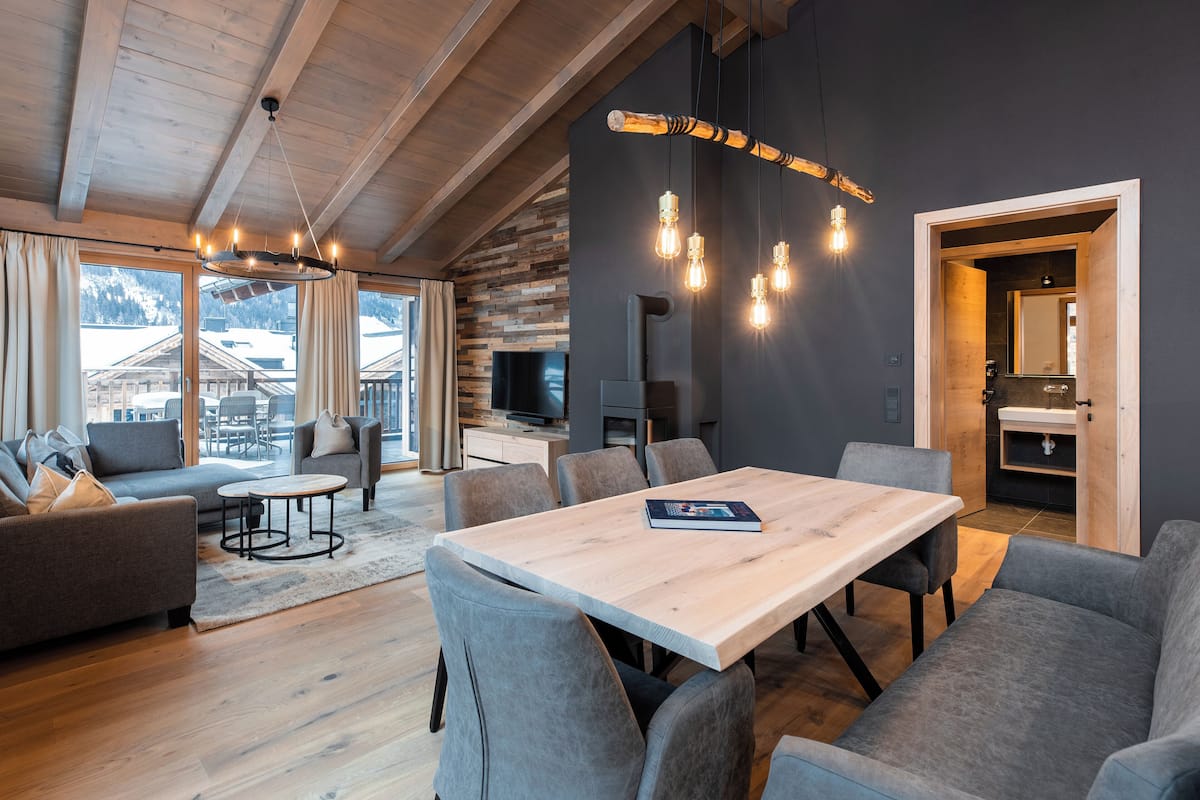 Een moderne woonkamer met een houten plafond, een eethoek met grijs gestoffeerde stoelen en een groot raam met uitzicht op een besneeerd landschap.