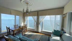 A dining area with a table and chairs, a chandelier, and large windows with curtains. - Assateague Point - Dynamic Waterfront ,Pier,Dock,  LINENS AND TOWELS PROVIDED (Chincoteague)