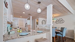 Una cocina con encimeras de granito, un lavamanos y taburetes de bar. Se observan lámparas colgantes de cristal y un espejo decorativo en la pared.