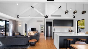 A spacious living area with a large sofa, a wooden coffee table, and a dining table with bar stools. The kitchen features a central island with bar stools and modern appliances. - Luxe Arrowtown Holiday Home (Arrowtown)