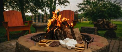 A circular fire pit with logs, surrounded by a stone fire ring. In front of the fire pit, there are marshmallows, chocolate bars, and cookies on a flat surface.