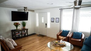 A living room with a wooden floor, a glass coffee table, leather sofas, a TV mounted on the wall, and a ceiling fan. - LAKEHOUSE/HOT TUB/8 MILES TO DOWNTOWN/ON 60 ACRES (Indianapolis)