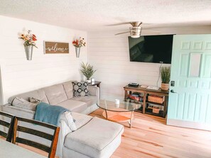 A living room with a sofa, a glass coffee table, a wooden side table, a television mounted on the wall, and a door with a light green color.