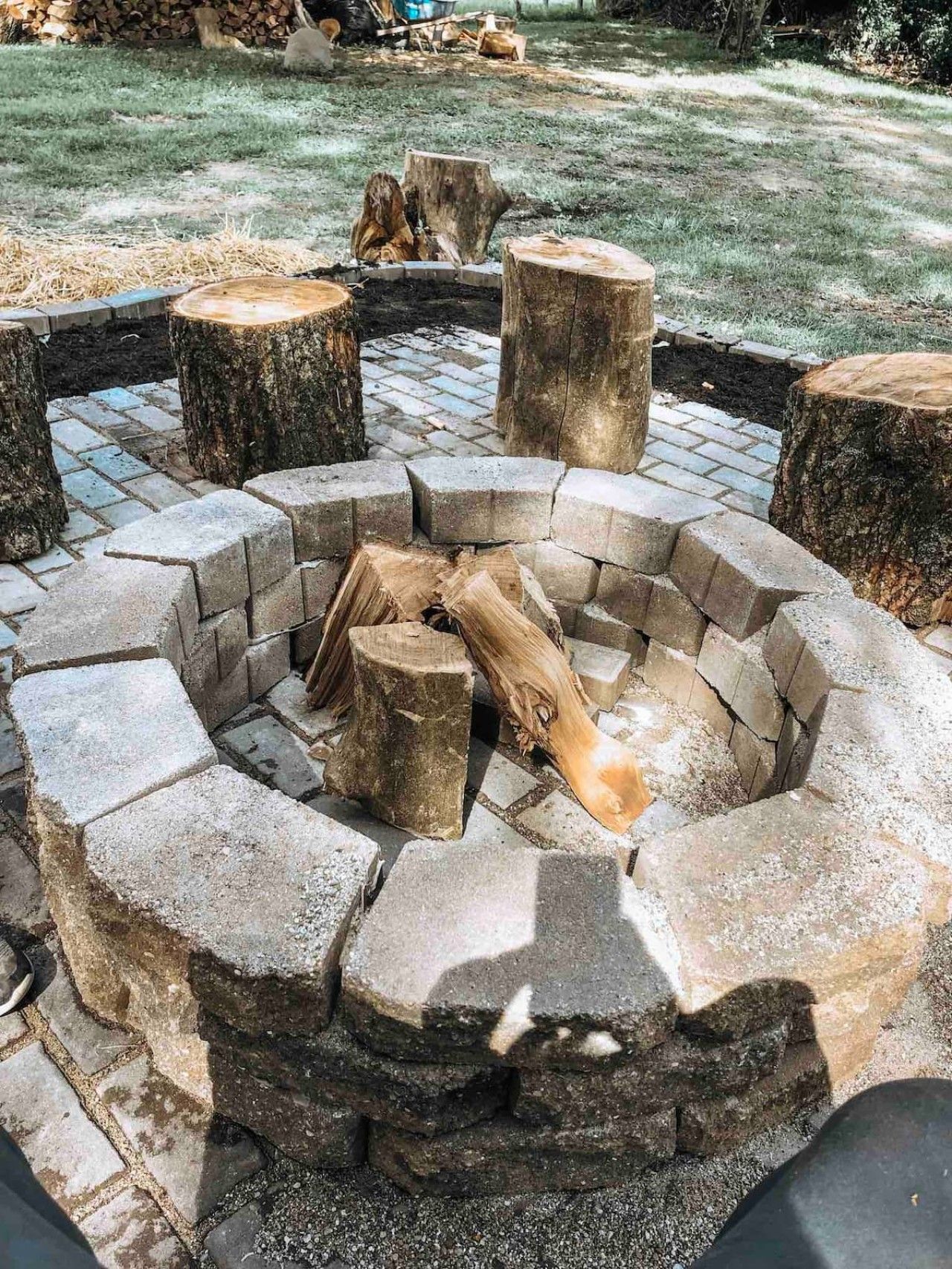 Un pozo de fuego circular de piedra con leña, rodeado por un borde de ladrillo, situado en un área verde al aire libre.