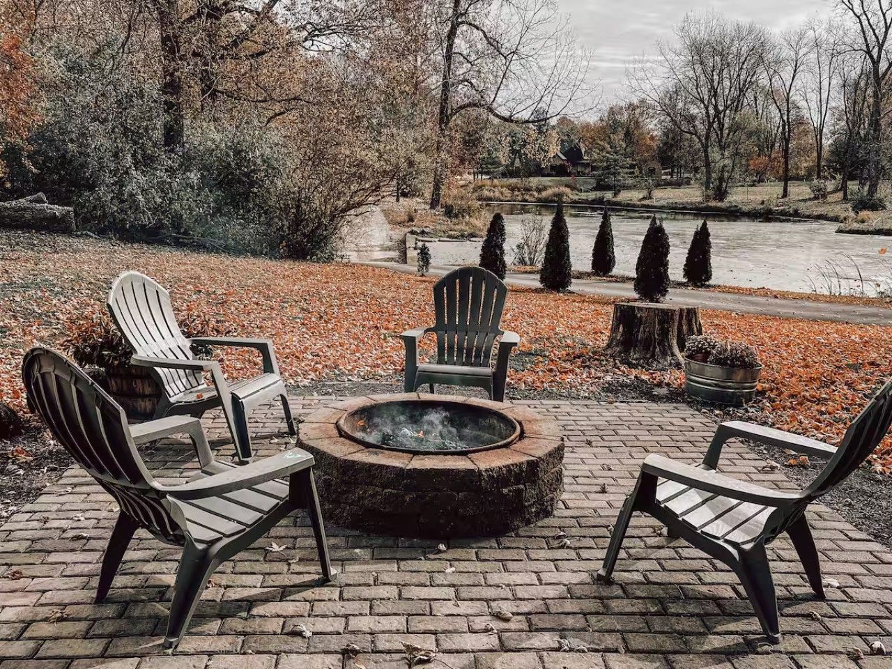A fire pit with four Adirondack chairs arranged around it, set on a brick patio with a view of a lake and trees in the background.