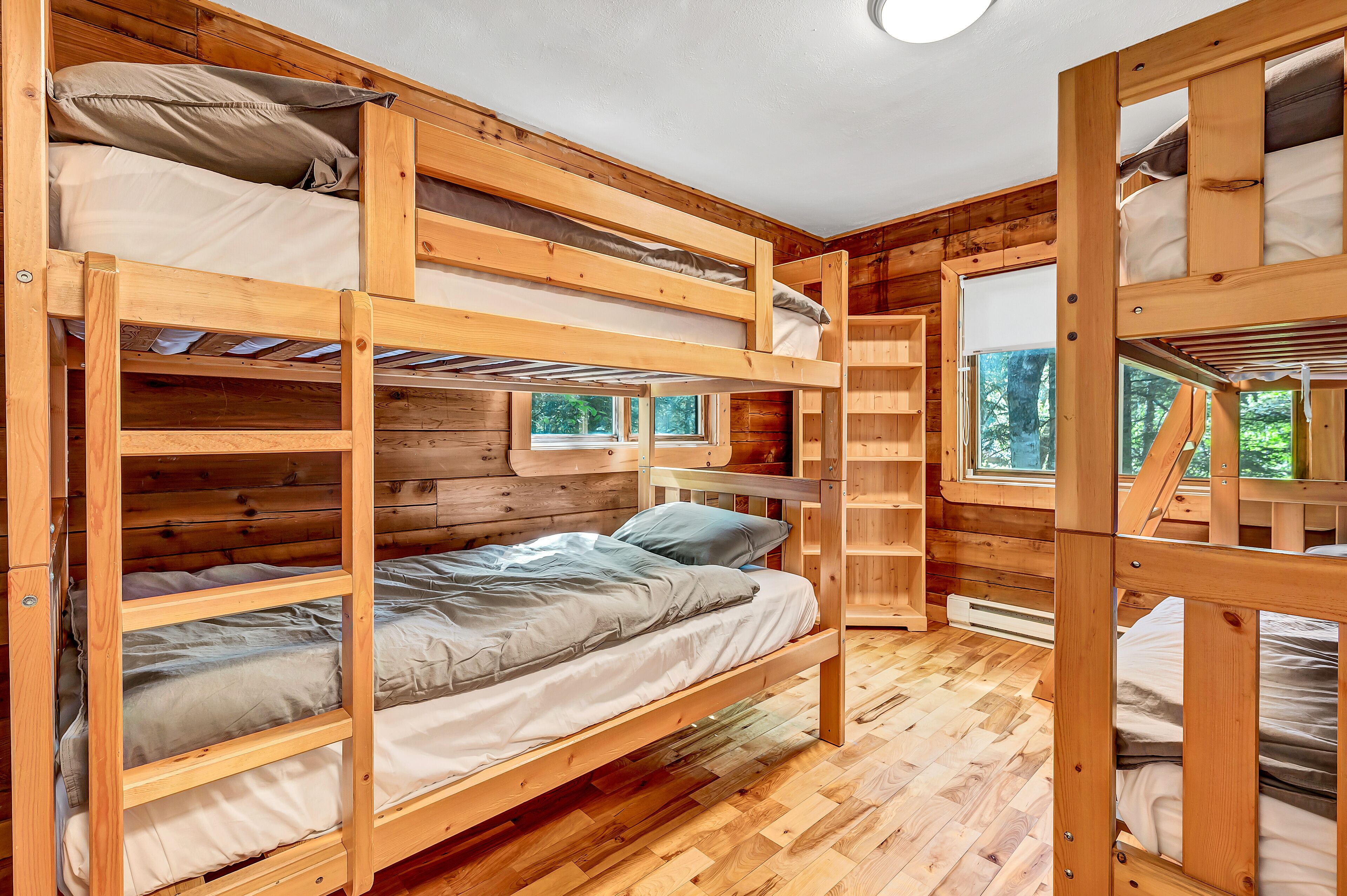 A bunk bed room with wooden walls and floors, a ladder, and a window with a view of trees.