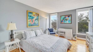 A bedroom with a bed, two chairs, a desk, and two paintings on the wall.
