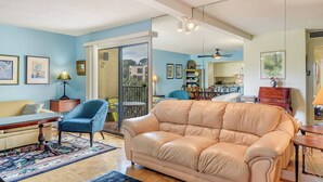 A living room with a beige sofa, a blue armchair, a yellow sofa, a dining table with chairs, and a kitchen area visible through a glass door.