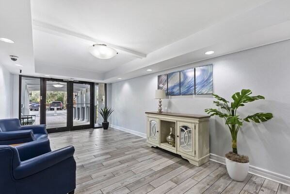 Una zona de recepción con piso de madera, sillas azules, un mueble con objetos decorativos y una planta grande en maceta.