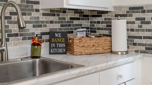 Una cocina con un lavamanos de acero inoxidable, un cartel que dice 'WE DANCE IN THIS KITCHEN', una cesta tejida con un paquete de café y un dispensador de toallas de papel blancas.