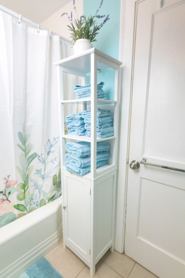 A bathroom with a white cabinet holding folded towels, a shower curtain with floral patterns, and a potted plant on the wall.