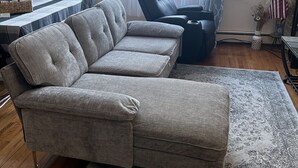 Una sala de estar con un sofá seccional gris, un sillón negro, una mesita con una lámpara y una ventana con cortinas traslúcidas.