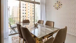 Un espace repas moderne avec une table en verre et quatre chaises, de grandes fenêtres offrant une vue sur les bâtiments et un élément décoratif mural.