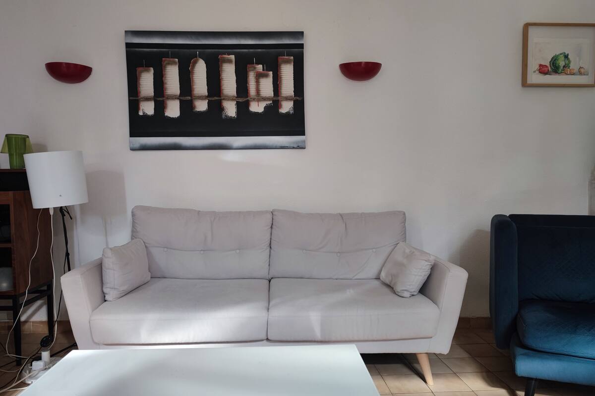 Uma sala de estar com um sofá branco, uma poltrona azul, uma mesa de centro branca, uma luminária e uma obra de arte emoldurada na parede.