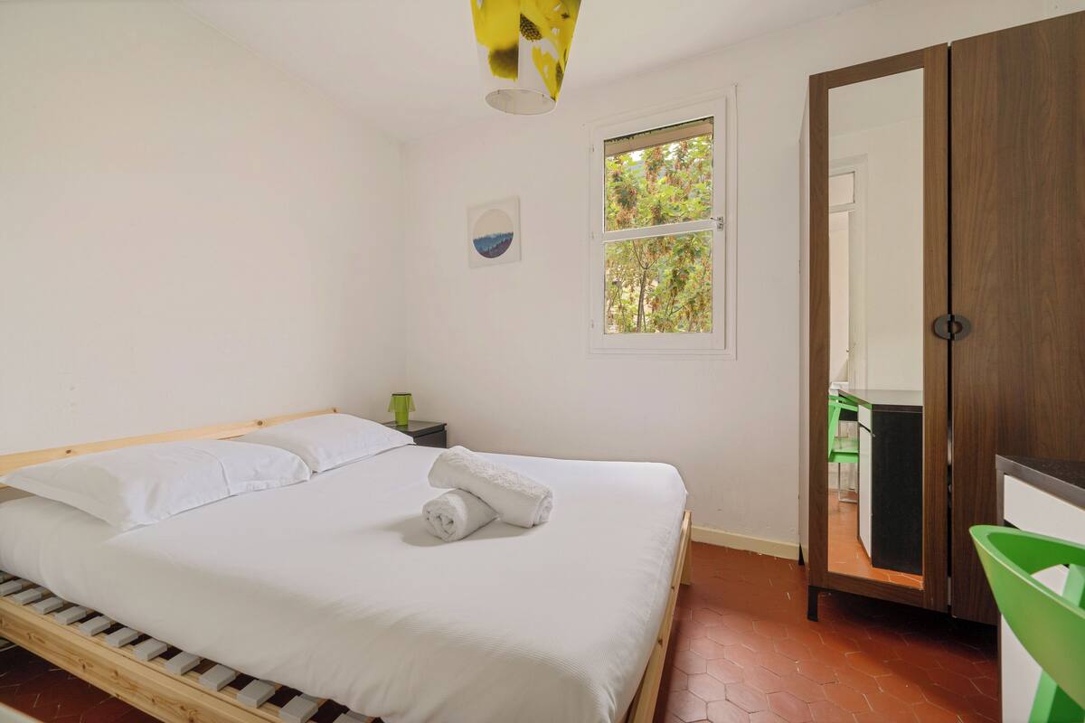 Cama bem arrumada com lençóis brancos e uma toalha enrolada, mesa de cabeceira pequena com abajur verde, janela com vista para árvores e guarda-roupa de madeira com espelho.