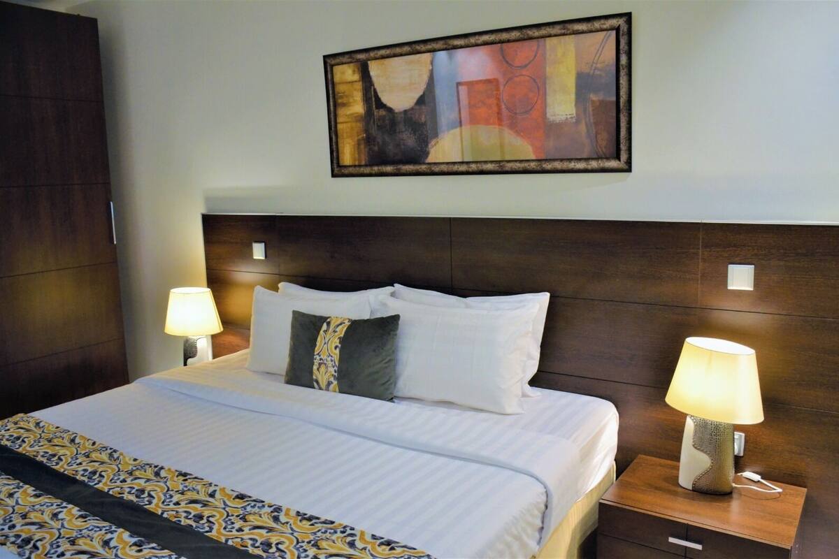 Una habitación de hotel con una cama grande, dos lámparas de noche, una mesita de noche y un cuadro abstracto enmarcado en la pared.