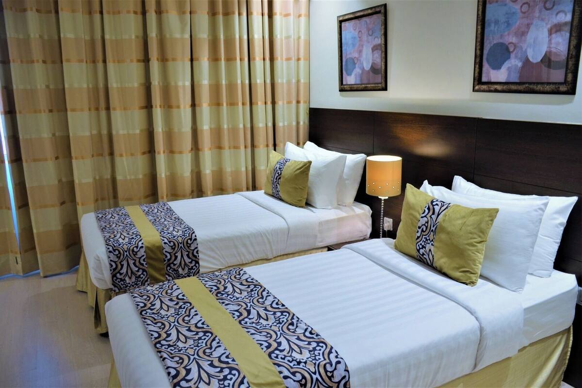 Una habitación de hotel con dos camas, cortinas color crema, cabecero de madera y una lámpara fijada a la pared.