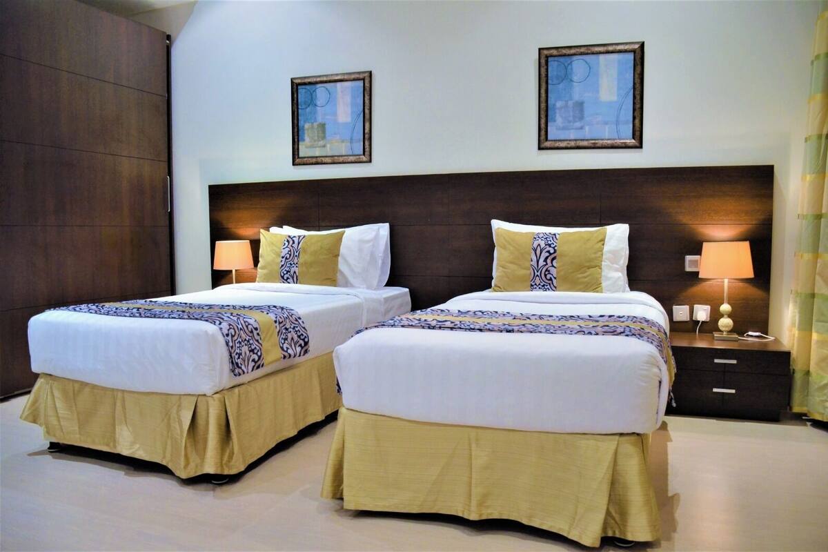 Habitación de hotel con dos camas, cada una con una colcha estampada y almohadas a juego. Hay dos mesitas de noche con lámparas, un cabecero grande de madera oscura y dos cuadros enmarcados en la pared.