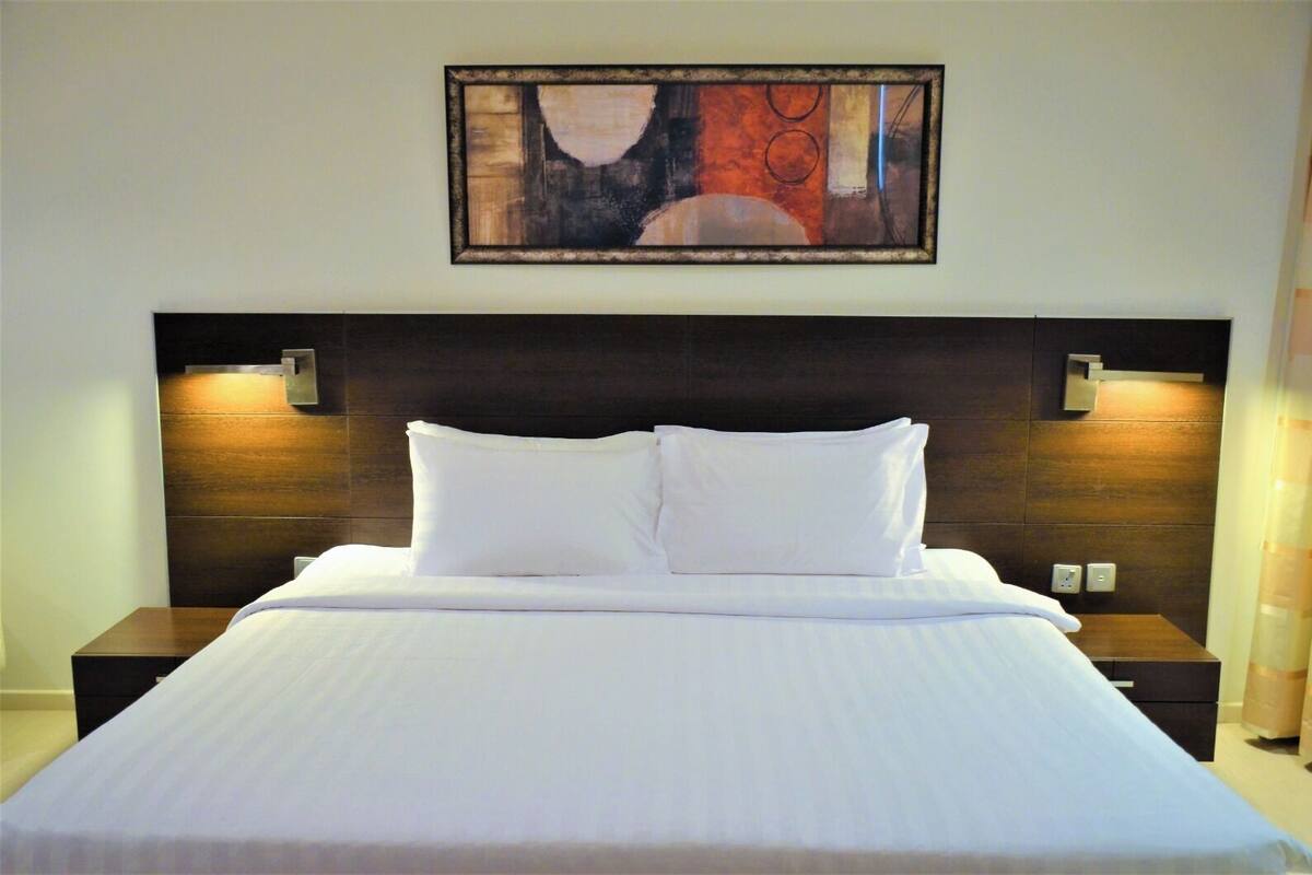 Una cama bien tendida con sábanas blancas, un cabecero de madera oscura y dos mesitas de noche con lámparas.